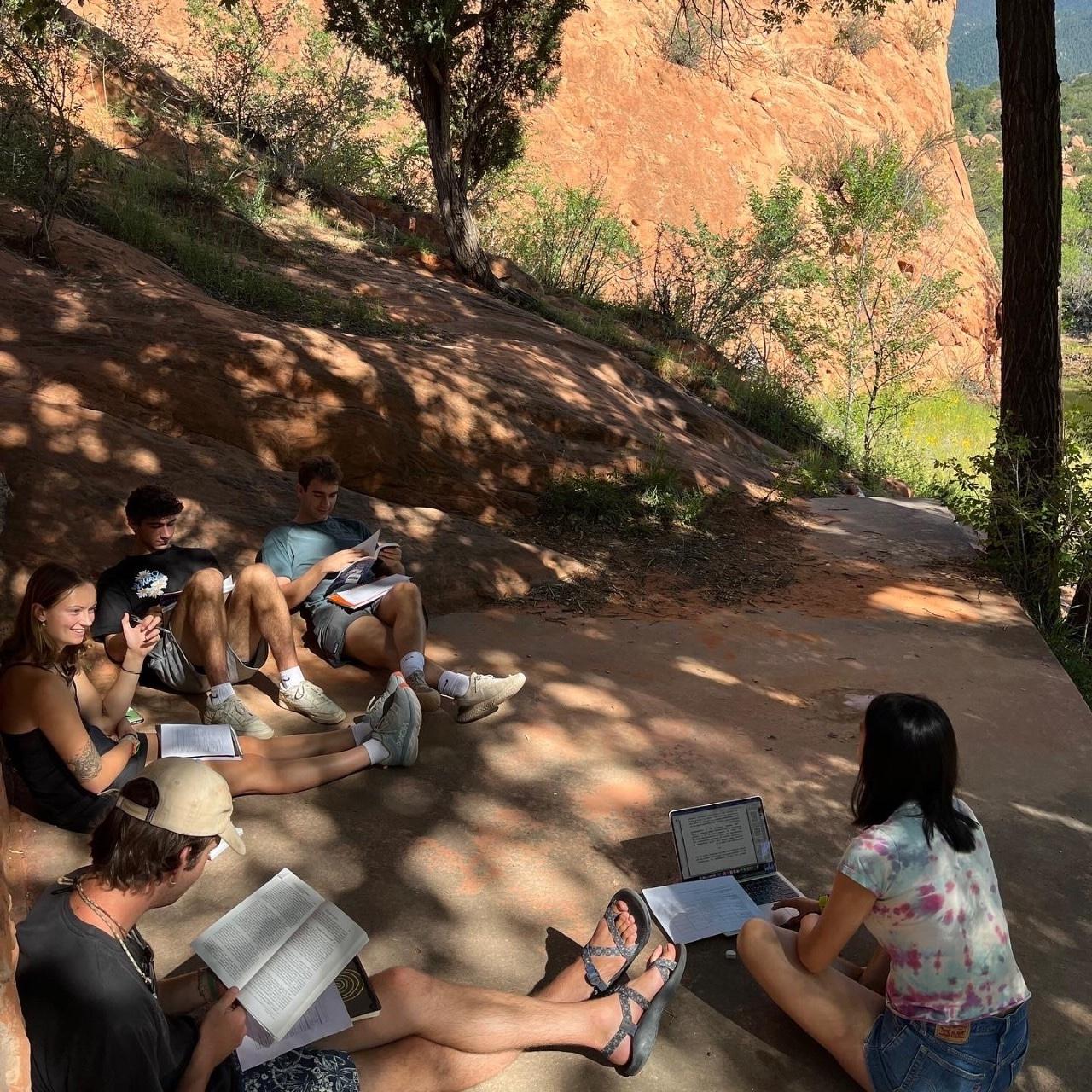 学生们拿着笔记本电脑和书本在红岩空地的树荫下讨论他们的阅读